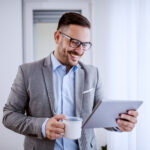 Attractive handsome classy caucasian smiling businessman in suit and with eyeglasses using tablet and holding mug with coffee while standing next to window in his office.